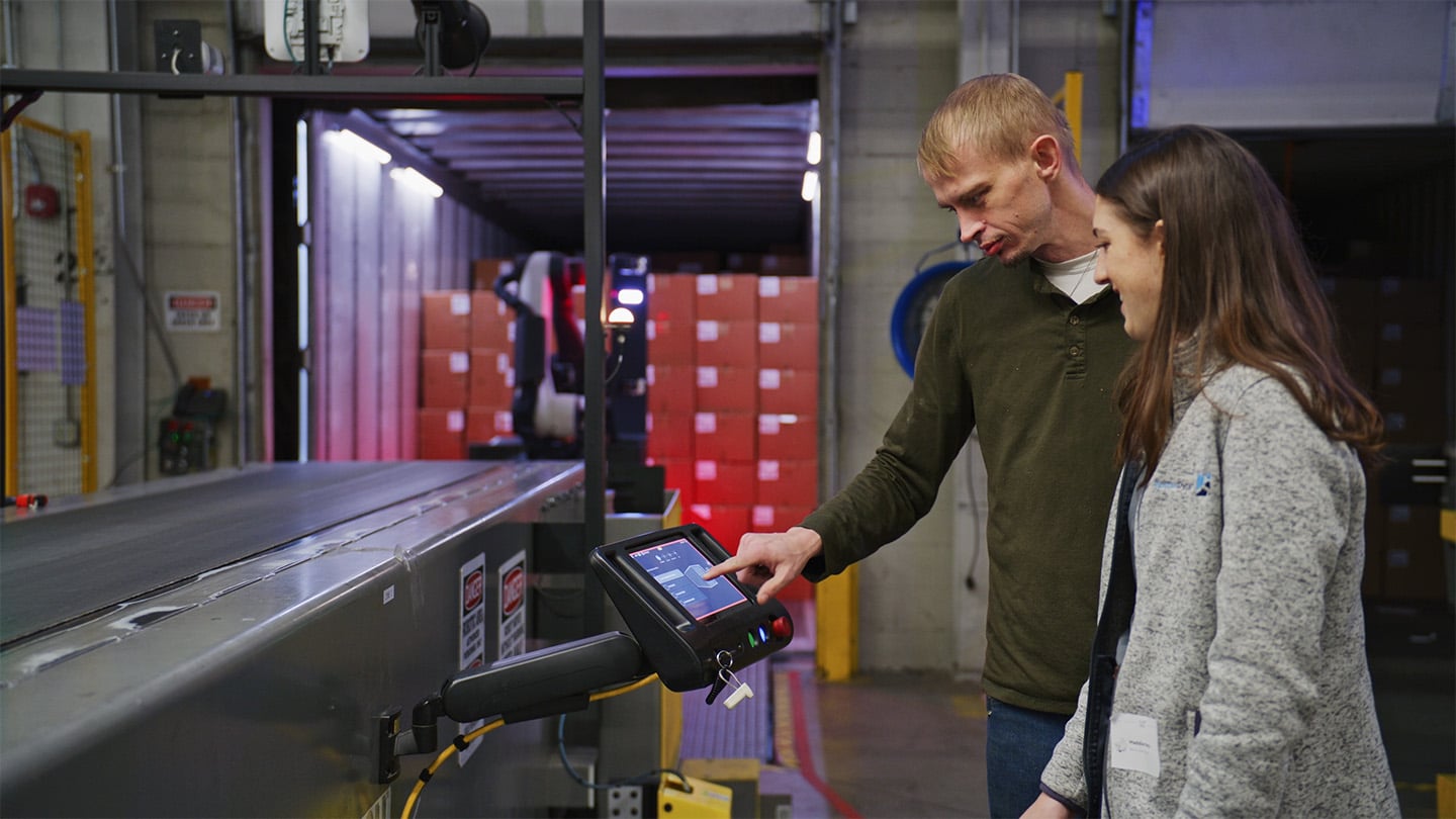 Warehouse worker touches console screen next to a Boston Dynamics engineer, with a conveyor, robot, and trailer in the background.